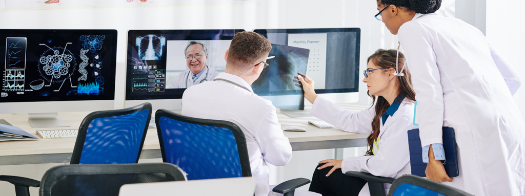 Three medical professionals in white lab coats are reviewing an X-ray film in a bright office. Two of them are seated in blue office chairs facing computer monitors displaying medical data and a video call with a doctor. The third person is standing, holding a tablet and observing the discussion. The environment suggests a collaborative medical consultation or telemedicine session.
