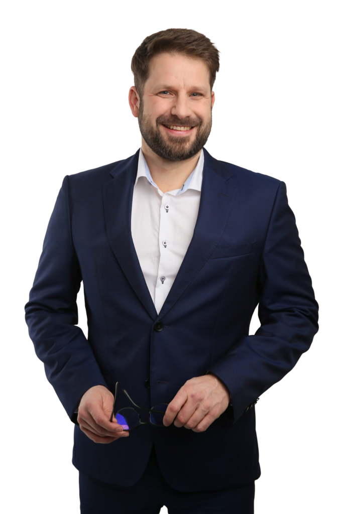 Man with short brown hair and beard wearing a dark blue suit and white dress shirt, smiling and holding black-framed glasses in both hands.